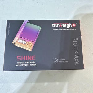May include: A rectangular black product box for a Truweigh Shine digital mini scale. The box features an image of a rainbow-colored scale with a chrome finish, displaying 0.00 on its screen. Text includes 100g x 0.01g, 10 Year Warranty, and Patent Pending.