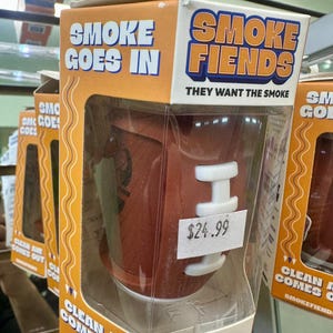 May include: A brown football-shaped air filter in a clear plastic case. The packaging reads "SMOKE FIENDS" and "SMOKE GOES IN, CLEAN AIR COMES OUT." The product is labeled "BLITZ, the football, eco-friendly personal air filter."
