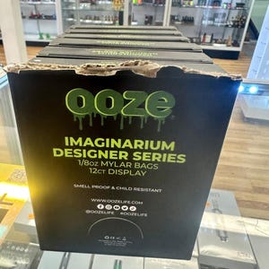 May include: A black display box with the word "ooze" in green dripping letters. The box reads "IMAGINARIUM DESIGNER SERIES 1/8oz MYLAR BAGS 12ct DISPLAY" and includes social media handles. The box is in a retail setting.