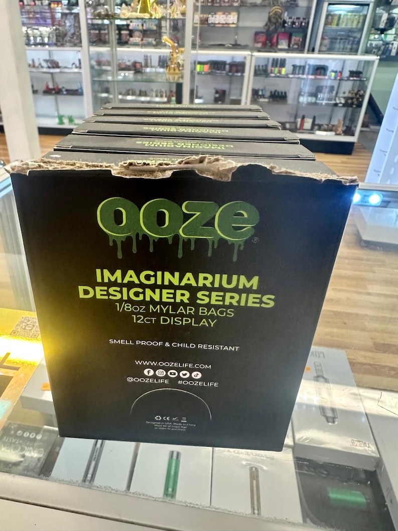 May include: A black display box with the word "ooze" in green dripping letters. The box reads "IMAGINARIUM DESIGNER SERIES 1/8oz MYLAR BAGS 12ct DISPLAY" and includes social media handles. The box is in a retail setting.