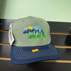 Könnte beinhalten: Grüne und marineblaue Baseballkappe mit einem gestickten Bären-Design und dem Wort "Utah". Der Bär ist mit einem Berg- und Sonnendesign dargestellt. Die Kappe hat einen Mesh-Rücken und einen gebogenen Schirm.