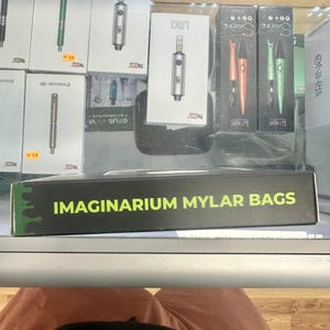 May include: A display of vaping products, including boxes labeled "LOKI" and "SARDINE", and pens in various colors. A box with the text "IMAGINARIUM MYLAR BAGS" is in the foreground. The products are arranged on a glass shelf.