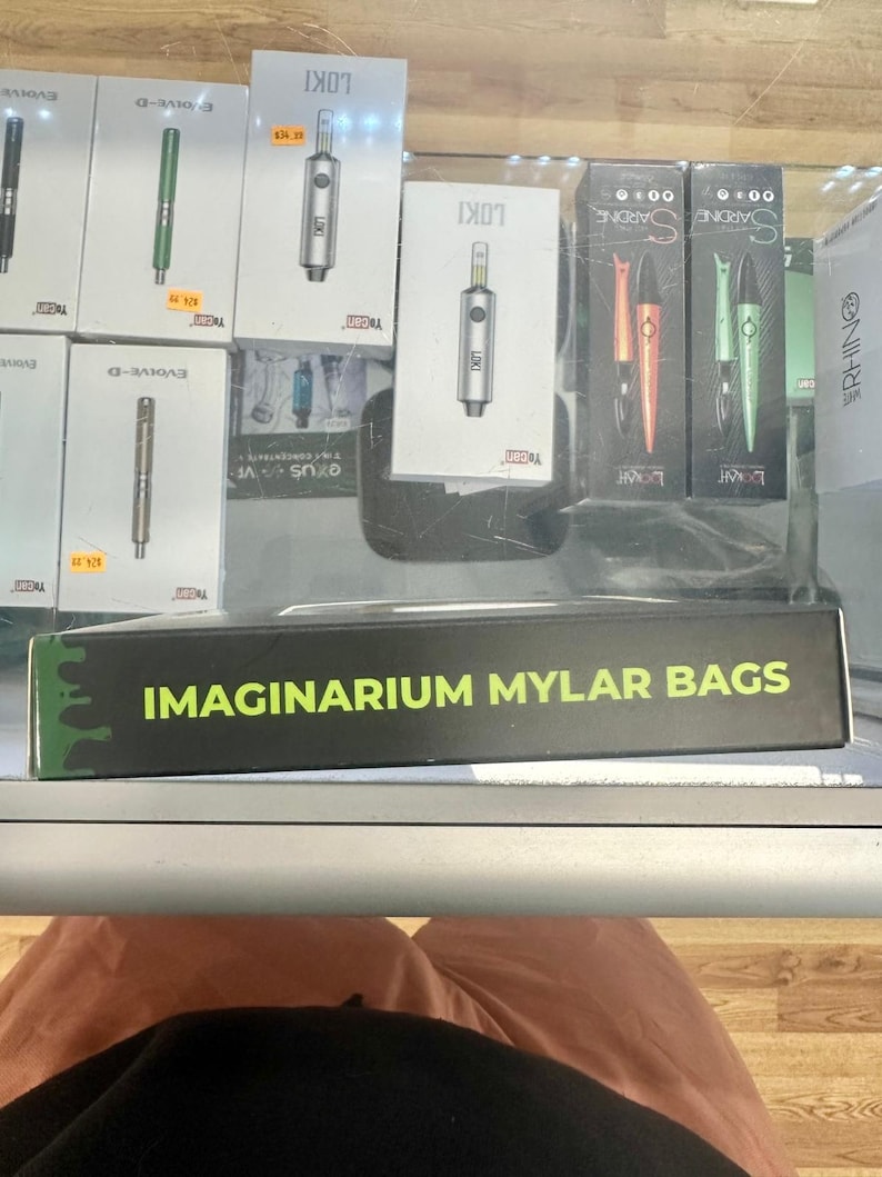 May include: A display of vaping products, including boxes labeled "LOKI" and "SARDINE", and pens in various colors. A box with the text "IMAGINARIUM MYLAR BAGS" is in the foreground. The products are arranged on a glass shelf.