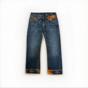 Puede incluir: Vaqueros azules con costuras decorativas y detalles coloridos. Los vaqueros presentan un diseño clásico de cinco bolsillos, con cierre de botón y cremallera. Los puños y los bolsillos delanteros tienen un diseño colorido.