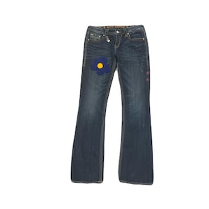 Könnte beinhalten: Dunkelblaue Jeans mit ausgestelltem Bein. Ein blauer Blumenaufnäher mit gelber Mitte befindet sich am linken Bein. Die Jeans hat dekorative Nähte und einen kleinen Anhänger an der Gürtelschlaufe.