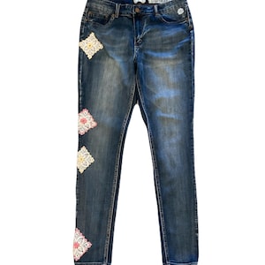 May include: Blue denim jeans with decorative crochet patches in cream, pink, and yellow. The jeans have a classic five-pocket design and a button and zipper closure. The jeans are a medium wash with slight fading.
