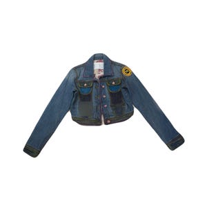 May include: A cropped denim jacket in shades of blue with patches of darker denim. The jacket has a button-up front, pink stitching, and a yellow patch on the upper sleeve. The jacket has two front pockets and two chest pockets.
