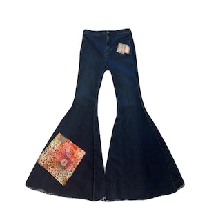 May include: Dark wash denim bell-bottom jeans with high waist. Features decorative patches on the front and back pockets. One large patch on the left leg has a colorful, patterned design. A stylish, retro-inspired garment.