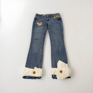 Può includere: Jeans di denim blu chiaro con una toppa a forma di cuore e ricami floreali. I jeans presentano inserti in tessuto bianco con dettagli di girasoli sugli orli svasati. Un design elegante e unico.