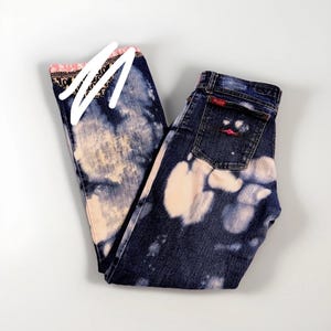 Peut inclure: Jean bleu foncé avec un motif blanchi. Le jean a une bordure rose et noire à motifs en haut des poches. La poche arrière a un logo rouge.