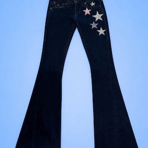 Puede incluir: Vaqueros de mezclilla azul oscuro con diseño de pierna acampanada. Los vaqueros presentan adornos de estrellas brillantes en rosa, plateado y morado en la parte delantera. Se muestran sobre un fondo azul claro.