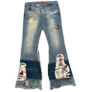 Puede incluir: Vaqueros de mezclilla azul claro con un diseño de pernera acampanada. Los vaqueros presentan parches bordados de un sol, un pato, un velero y una mujer con una bolsa de la compra. El dobladillo inferior tiene un lavado azul más oscuro.