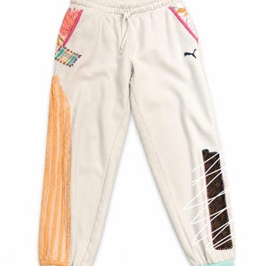 May include: Cream-colored sweatpants with colorful patchwork details. Features an orange textured stripe, a small Puma logo, and a drawstring waist. The cuffs are light blue, and the patches include pink, brown, and patterned fabrics.