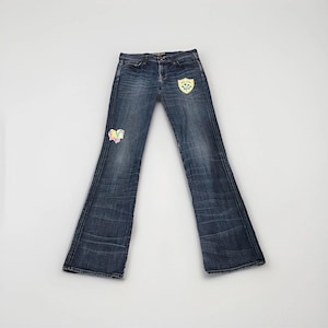 May include: Dark wash denim jeans with a flared leg. The jeans feature decorative patches on the front, including a heart and a shield-shaped design. The jeans have a classic five-pocket design and a button and zipper closure.