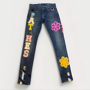 May include: Blue denim jeans with colourful embroidered patches and floral designs. The word "PATCHES" is vertically embroidered on the left leg. Features a butterfly patch and a yellow flower patch.