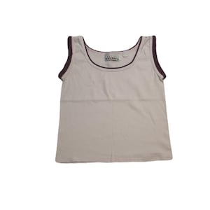 May include: A light beige tank top with a scoop neck and dark burgundy trim. The tank top has a small tag near the neckline with text. The sleeveless top is made of a soft material.