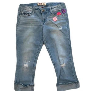 May include: Light blue denim capri pants with floral embroidery and distressed details. Features a button and zipper closure, front and back pockets, and rolled cuffs. The brand label "lei" is visible on the waistband.