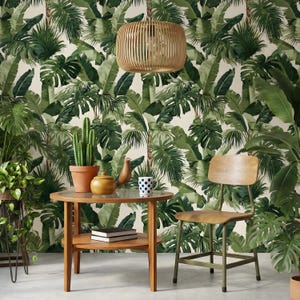 Puede incluir: Una escena interior de temática tropical con una mesa de madera, una silla de madera y una lámpara colgante tejida. El fondo es un papel pintado con exuberante follaje verde. Un cactus en maceta y una taza con lunares azules están sobre la mesa.