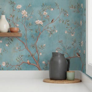 Puede incluir: Un mural de pared de color turquesa con un diseño floral y de pájaros. El mural representa ramas con flores blancas y pájaros pequeños. Una jarra gris, una pequeña taza verde y una bandeja de madera están en primer plano.