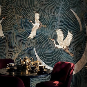 Puede incluir: Una escena de comedor con una mesa redonda oscura con un juego de té dorado y tazas de té negras. El fondo es un papel pintado verde azulado y dorado con grullas volando. Se ven dos sillas de color burdeos.