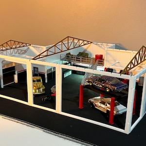 Könnte beinhalten: Miniatur-Diorama einer Garage mit weißer Außenfassade, braunen Dachsparren und roten Akzenten. Zwei Spielzeugautos stehen auf einer Hebebühne, ein weiteres ist geparkt. Die Szene umfasst einen kleinen LKW und eine Plattform.