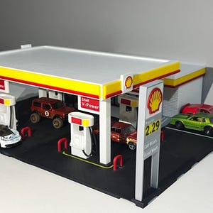 May include: A miniature Shell gas station model with a white and yellow canopy. The model includes several toy cars, gas pumps, and signage. The gas station has a black base and red accents.