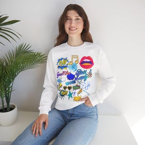 Könnte beinhalten: Weißes Sweatshirt mit farbenfrohen Pop-Art-Grafiken. Das Design umfasst Sprechblasen mit Wörtern wie "POW" und "WOW", Noten und Lippen. Das Sweatshirt wird mit einer Jeans getragen.