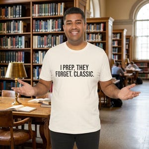 Op de afbeelding: Een crèmekleurig t-shirt met de tekst "I PREP, THEY FORGET. CLASSIC." in het zwart. Het shirt wordt gedragen door een persoon in een bibliotheek, met boekenkasten vol boeken op de achtergrond.