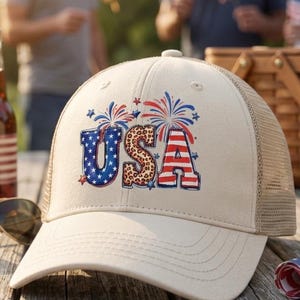 以下が含まれることがあります： 赤、白、青で「USA」の文字と花火が描かれたベージュ色のトラッカーハット。 サングラス、アメリカ国旗のラベルが付いたボトル、丸められたアメリカ国旗が木製のピクニックテーブルに置かれています。