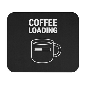 May include: Black square mouse mat featuring the white text "COFFEE LOADING" above a white outline of a coffee mug with a loading bar. The mug has a handle on the right. The design is simple and modern.