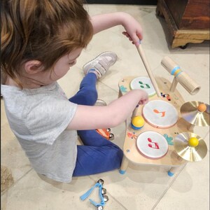 May include: A child's wooden toy drum set with a light wood finish. The set includes three drums with musical note designs, two cymbals, a yellow ball, and a wooden microphone. A pair of drumsticks are held by the child.