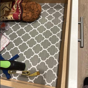 May include: An open wooden drawer with a gray and white patterned liner. A brown and red oven mitt, a blue marker, a black marker, and a yellow measuring tape are visible inside the drawer.