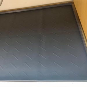 May include: A gray, rectangular shelf liner with a repeating chevron pattern. The liner is inside a wooden cabinet, with a white edge and a metal hinge visible on the right side. The liner is designed to protect the shelf.