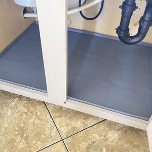 May include: A gray, hexagonal-patterned sink cabinet mat is installed inside a light-colored cabinet. Plumbing pipes and a white water supply line are visible above the mat. The cabinet sits on a tiled floor.