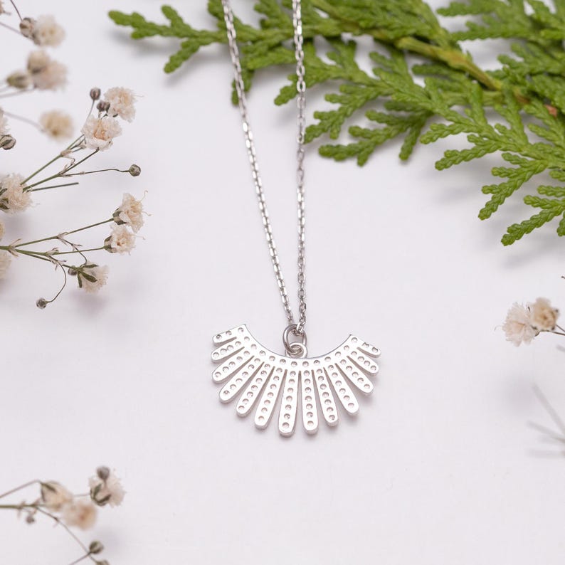 May include: A silver necklace featuring a fan-shaped pendant with a series of small, evenly spaced cutouts. The necklace is displayed on a white surface, adorned with sprigs of greenery and small white flowers. The pendant is a decorative piece.