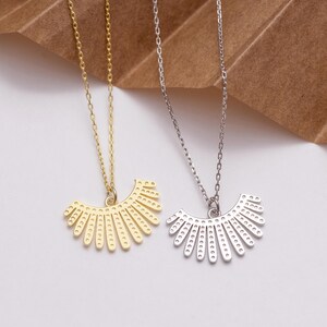 May include: Two necklaces with fan-shaped pendants. One is gold, the other silver. Both pendants feature small, evenly spaced holes along the curved edge and are suspended from delicate chains.