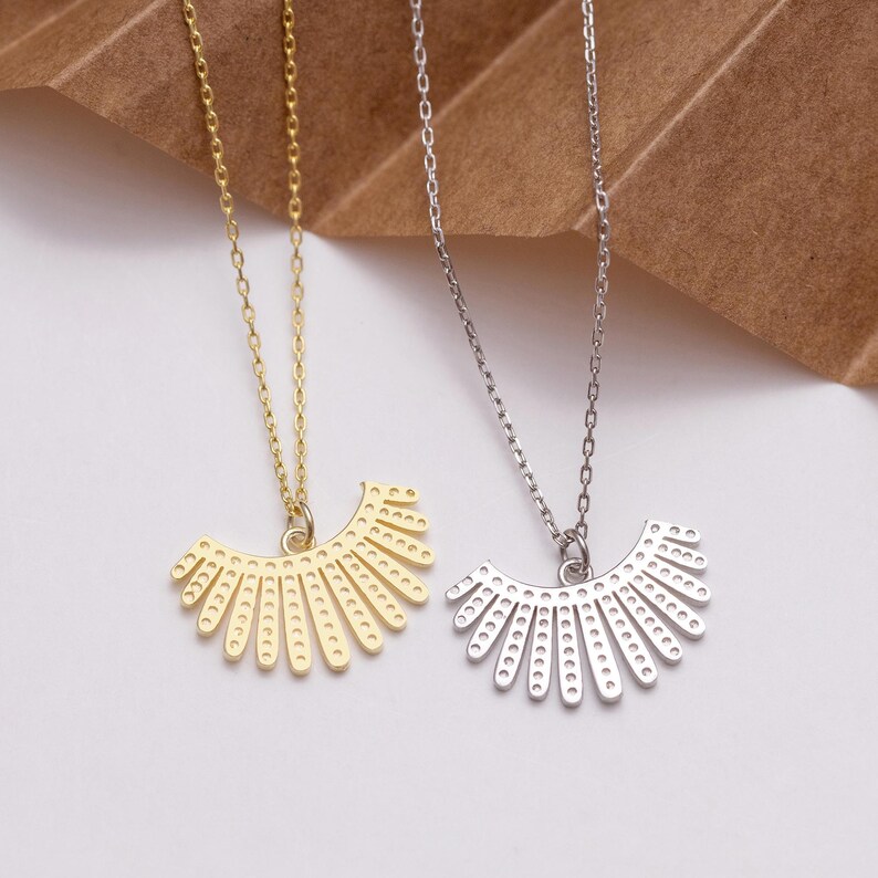 May include: Two necklaces with fan-shaped pendants. One is gold, the other silver. Both pendants feature small, evenly spaced holes along the curved edge and are suspended from delicate chains.