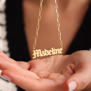 May include: A gold-toned necklace featuring a nameplate pendant with the name Madeline written in a stylized gothic font. The pendant is attached to a delicate paperclip chain, which consists of elongated, rectangular, interlocking gold links.