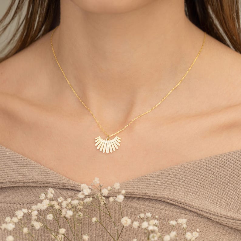 May include: A gold necklace with a sunburst pendant. The pendant has a fan-shaped design with vertical lines. The necklace is worn, and the background is blurred. The necklace is a delicate piece of jewellery.