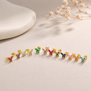 May include: A collection of gold stud earrings shaped like butterflies, each featuring small, colorful gemstones in shades of red, purple, light blue, clear, green, pink, dark blue, and orange, arranged in a horizontal row on a flat surface.