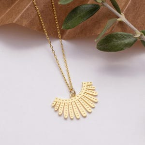 May include: A gold necklace featuring a sunburst pendant. The pendant has a series of curved lines with small dot details. The necklace chain is delicate and gold. The background is neutral with a hint of greenery, with a brown paper element.