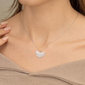 May include: A silver necklace with a fan-shaped pendant set with small, sparkling stones. The pendant is suspended from a delicate chain, creating a simple, elegant design. The necklace is displayed against a neutral backdrop.