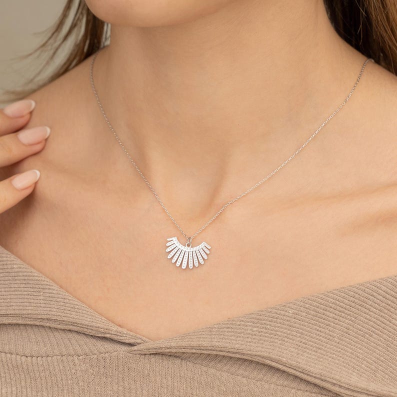 May include: A silver necklace with a fan-shaped pendant set with small, sparkling stones. The pendant is suspended from a delicate chain, creating a simple, elegant design. The necklace is displayed against a neutral backdrop.