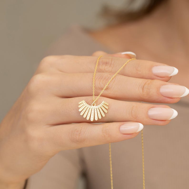 May include: A gold necklace with a sunburst pendant. The pendant has a half-circle design with vertical lines and small, sparkling details. The necklace is shown against a neutral background.