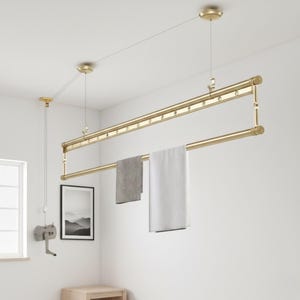 Może przedstawiać: Złoty, sufitowy wieszak na pranie z dwoma poziomymi drążkami. Wieszak jest zawieszony na drutach i posiada system bloczkowy. Dwa ręczniki, jeden szary i jeden biały, wiszą na drążkach. Pokój jest jasny i biały.