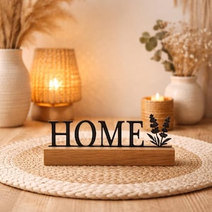 Peut inclure: Un panneau en bois avec le mot 'HOME' en noir et un motif floral. Le panneau est posé sur une base en bois clair, sur un tapis tissé. L'arrière-plan comprend des vases avec des plantes séchées et une bougie allumée.