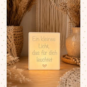 Op de afbeelding: Een rechthoekige witte lichtbak met de Duitse tekst "Ein kleines Licht, das für dich leuchtet" en een klein hartpictogram onderaan. Het apparaat verspreidt een zachte, warme gloed van binnenuit, waardoor de tekst duidelijk verlicht wordt.
