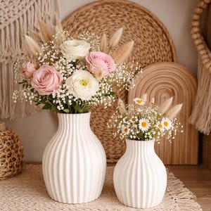 Könnte beinhalten: Zwei weiße, geriffelte Keramikvasen mit frischen Blumen. Die größere Vase enthält rosa und weiße Rosen, die kleinere Gänseblümchen. Die Vasen stehen auf einem Holzregal, mit gewebten Dekorationen im Hintergrund.
