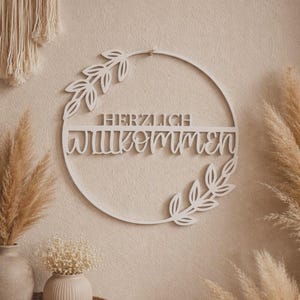 Könnte beinhalten: Ein weißes, kreisförmiges Holzschild mit den deutschen Worten "HERZLICH WILLKOMMEN" (Willkommen) ausgeschnitten. Das Schild ist mit Blattmotiven verziert und hängt an einer beigefarbenen Wand.