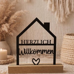 Könnte beinhalten: Schwarzes, lasergeschnittenes Holzschild in Hausform mit den Worten "HERZLICH Willkommen" und einem Herzsymbol. Das Schild ist auf einem Naturholzsockel montiert. Der Hintergrund umfasst eine Kerze, Trockenpflanzen und einen Weidenkorb.
